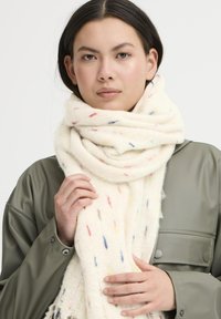 Krémfarvet uld scarf med farverige prikker, blød tekstur og frynsetender. Modellen har en grå frakke på og holder blidt om tørklædet.