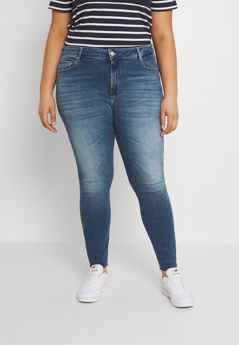 Person i bleknade blå skinny jeans, vita sneakers och en svart-vitrandig kortärmad skjorta står mot en vit bakgrund.