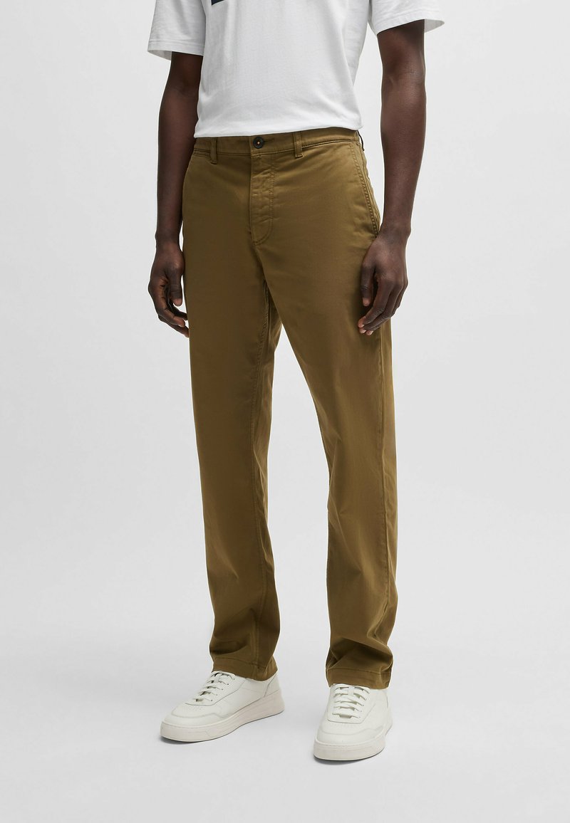 Khaki katoenen broek met rechte pijpen, voorzien van een knoopsluiting aan de voorkant en zijzakken, gedragen met witte sneakers.