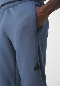 Niebieskie spodnie sportowe z gładkiego materiału, z bocznymi kieszeniami i czarnym logo Adidas na dolnej lewej nogawce.