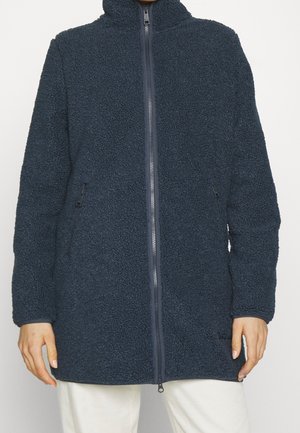 Persona che indossa una giacca in pile blu scuro con cerniera integrale, tasche laterali e collo alto, abbinata a pantaloni bianchi.