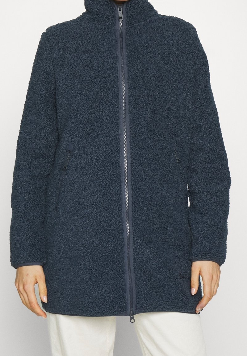 Personne portant une veste polaire bleu foncé zippée intégrale avec poches latérales et col montant, associée à un pantalon blanc.