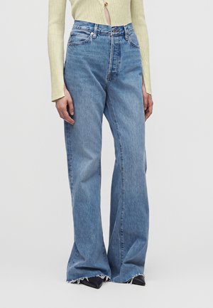 Person iført lyseblå vide jeans med flossede kanter og sorte spidse sko, kombineret med en lysegul ribbet bluse med lange ærmer.