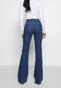 Högmidjade utsvängda jeans i mörkblå denim med två bakfickor och subtila sömdetaljer. Slät textur med en åtsittande midja.