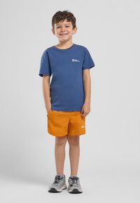 Blaues T-Shirt mit kurzen Ärmeln und Logo, kombiniert mit leuchtend orangefarbenen Shorts und grauen Schuhen mit schwarzen Sohlen und Akzenten. Geeignet für aktive Kleidung.