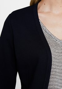 Cardigan in maglione blu navy con scollatura a V, indossato sopra una maglietta a righe bianco e nere. Tessuto liscio con sottili dettagli a coste.