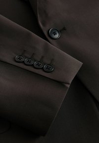 Brauner Anzugsjackenärmel mit glattem Stoff, vier schwarzen Knöpfen und detaillierter Nahtverzierung in der Nähe der Manschetten.