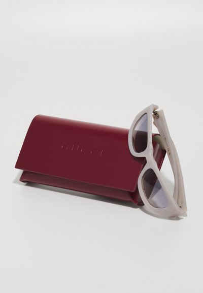 Slnečné okuliare Gucci s matným bielym rámom a sivými sklami, umiestnené vedľa vínovo červenej koženej etui s vyrazeným nápisom "Gucci."
