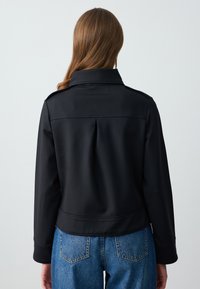 Zwarte jas met een hoge kraag, voorzien van een rugplooien en een gladde textuur. Gecombineerd met blauwe hoge taille jeans, die een casual pasvorm laten zien.