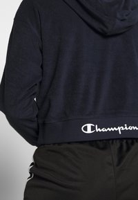 Felpa con cappuccio blu marina realizzata in tessuto morbido, con un taglio cropped e un logo "Champion" bianco sulla cintura a coste. Abbinata a pantaloni neri.