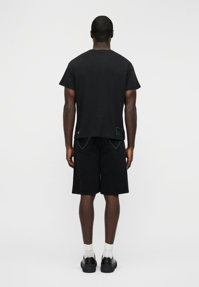 Μαύρο βαμβακερό t-shirt με άνετη εφαρμογή, συνδυασμένο με μαύρα τζην σορτς. Εμφανείς μοβ λεπτομέρειες κοντά στο τελείωμα. Λευκές κάλτσες και μαύρα παπούτσια.