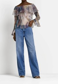 Sheer, multicolored floral blouse with layered ruffles, off-the-shoulder design, paired with wide-leg blue jeans and brown sandals.