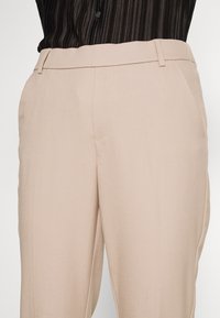 Pantalon beige ajusté avec une texture lisse, doté d'un devant plat, de passants de ceinture et de poches latérales. Le tissu a un léger éclat.