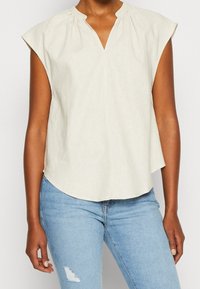 Top sin mangas de color beige claro con escote en V y detalles fruncidos, hecho de una tela suave. Combinado con jeans azul desgastado con una rodilla desgastada.