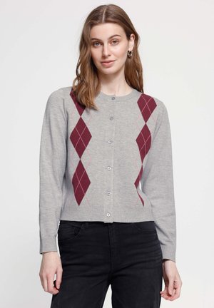Femme portant un cardigan gris boutonné avec un motif losanges bordeaux et un pantalon noir, debout devant un fond clair uni.