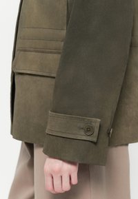 Casaco de camurça verde-oliva com um design estruturado, punhos com botões e bolsos laterais. O material tem uma textura suave e lisa.
