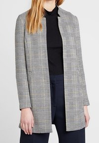 Abrigo de cuadros gris con collar mandarín, que presenta un corte holgado, mangas largas y bolsillos laterales. Está hecho de una tela texturada con patrones de cuadros en blanco y negro.