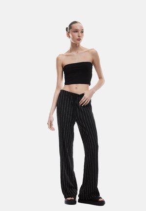 Zwarte geribbelde strapless top in combinatie met losse zwarte broeken met witte verticale strepen. De outfit wordt aangevuld met slanke zwarte slides.