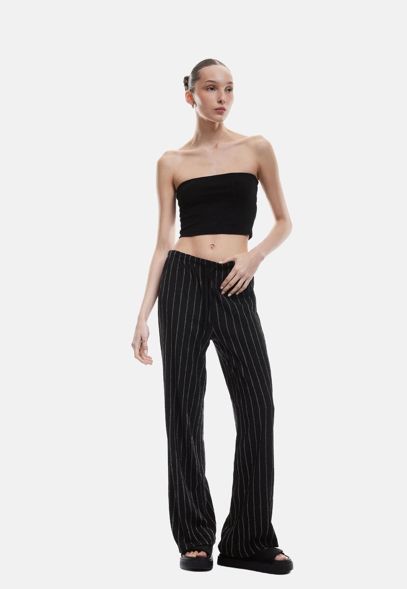 Schwarzes, geripptes, trägerloses Top, kombiniert mit lockeren schwarzen Hosen mit weißen vertikalen Streifen. Das Outfit wird durch elegante schwarze Slides ergänzt.