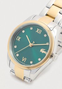 Montre-bracelet en argent et or avec un cadran vert, une lunette texturée, des index horaires en or et de petits cristaux ; comprend un logo de crocodile en bas.