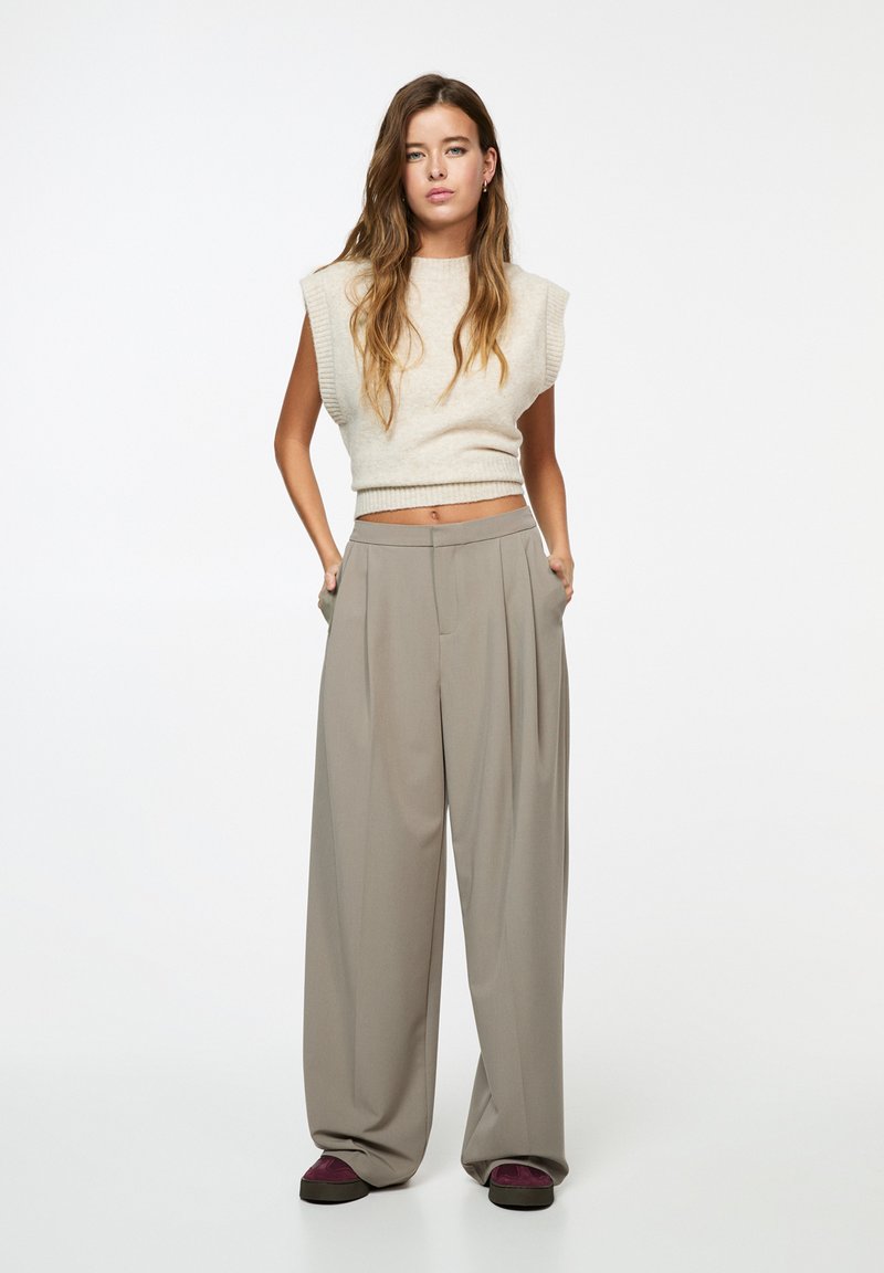 Haut en tricot sans manches beige associé à un pantalon large à taille haute plissé taupe. Comprend des poches latérales et des chaussures foncées avec une touche de violet.