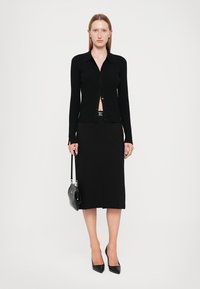 Black ribbed long-sleeve cardigan with buttons, paired with a black knee-length skirt. Accessories include a small black bag and pointed heels.