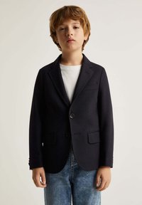 Blazer navy de tela texturada, con cierre de dos botones, solapas en pico y dos bolsillos frontales, llevado sobre una camisa blanca.