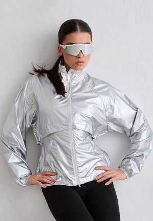 Kobieta w błyszczącej srebrnej kurtce na zamek i refleksyjnych sportowych okularach przeciwsłonecznych, pozująca z rękami na biodrach na tle jednolitej ściany.