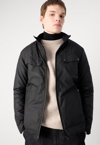 Schwarze jacke mit Reißverschluss, großen Fronttaschen, Knopfakzenten und Stehkragen, getragen über einem beigen gerippten Pullover.