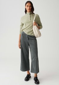Femme portant un pull léger vert, un pantalon large gris, des chaussures noires et tenant un sac à épaule couleur crème.