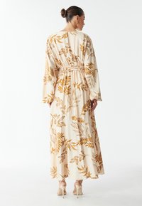 Vrouw met een haar in een knot draagt een crème maxi-jurk met lange mouwen en een bruin bladdessin, staand met haar rug naar de kijker, beige hakken zijn zichtbaar.