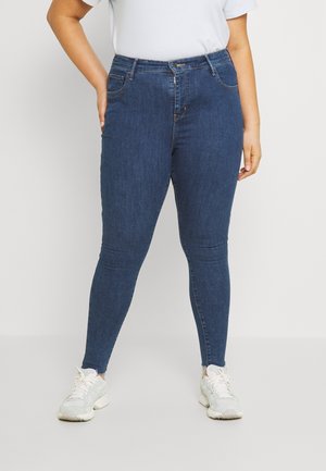 Personne portant un jean skinny bleu foncé et des baskets blanches, debout avec une main posée sur la hanche.