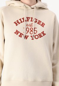 Felpa color crema in materiale morbido con cappuccio regolabile. Presenta un testo ricamato in rosso che dice "HILFIGER est 1985 NEW YORK" sulla parte anteriore.