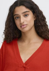 Cardigan rosso lavorato a maglia con scollo a V profondo e chiusura con fiocco, caratterizzato da una texture a coste lungo i bordi e una finitura morbida e soffice.