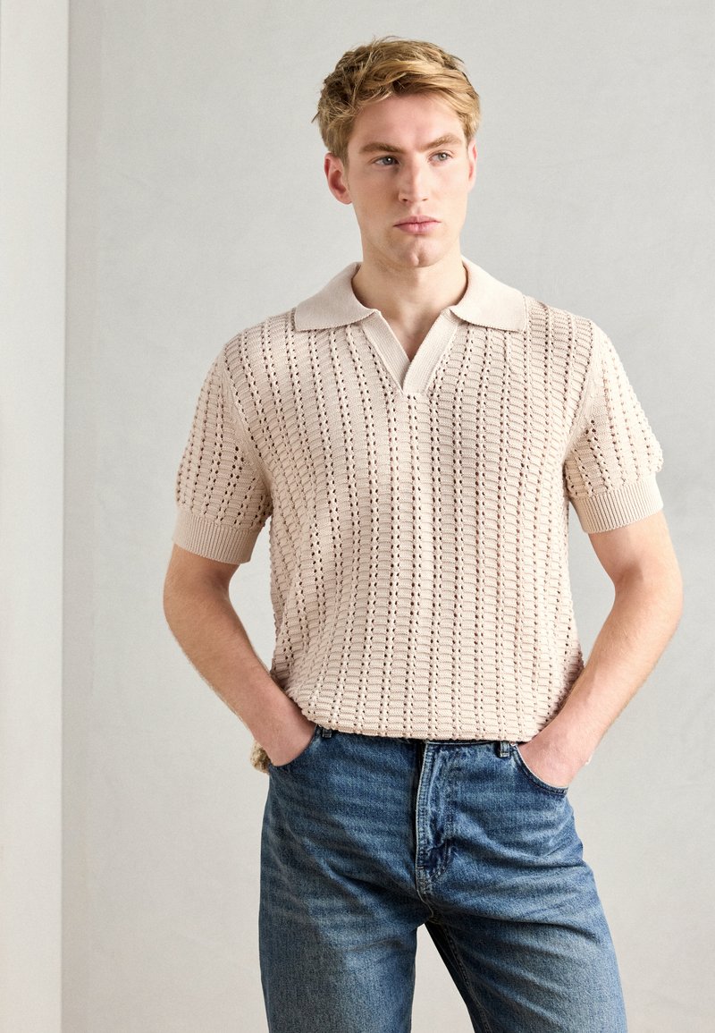 Beige knitted polo shirt with a v-neck, short sleeves, and a ribbed collar. Features a textured pattern with an open weave. Worn with blue jeans.