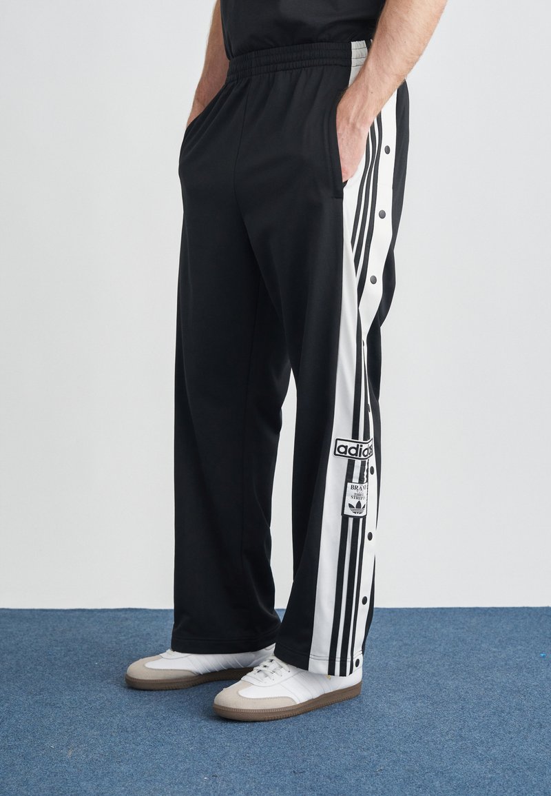 Čierne športové nohavice s detailmi bielych patentných gombíkov a tromi vertikálnymi pruhmi. Na ľavej strane je logo Adidas.