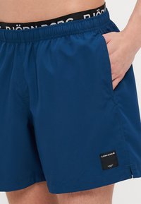 Navy blue swim shorts with an elastic waistband, side pocket, and a black tag featuring white logo. Smooth fabric with a casual fit.