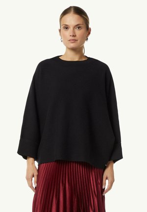 Pull en tricot noir surdimensionné avec un col rond et des manches larges, porté sur une jupe rouge plissée, mettant en avant une texture douce et une coupe décontractée.