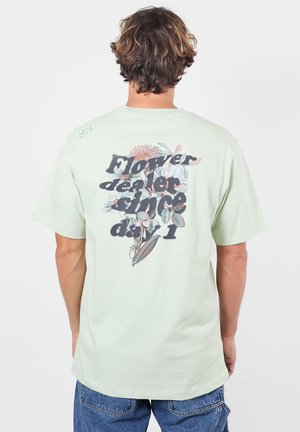 Personne aux cheveux courts et bruns porte un t-shirt vert clair avec un motif floral et le texte "Flower dealer since day 1" dans le dos.
