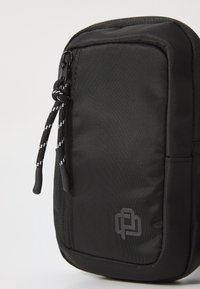 Pochette nera in tessuto con chiusura a zip, superficie testurizzata e accentuazione del logo. Presenta una corda per il tiraggio con strisce bianche a contrasto.
