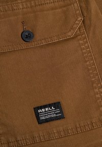 Brown corduroy fabric with vertical ribbing, featuring a black button and a label that reads "REELL ESTABLISHED 1997."