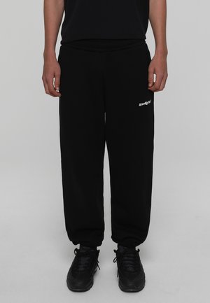 Personne portant un pantalon de jogging noir avec un logo blanc « lowlight » sur la cuisse et des baskets noires, debout devant un fond gris uni.