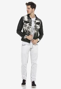 Herrenjacke mit einem schwarzen Lederunterteil und einem gemusterten weißen Stoffoberteil. Kombiniert mit weißen Jeans und schwarzen Sneakers.