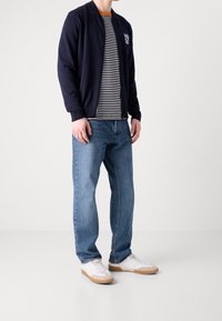 Marinblå bomberjacka över en randig T-shirt, parat med blå jeans och vita sneakers med beige detaljer. Klassisk avslappnad outfit.