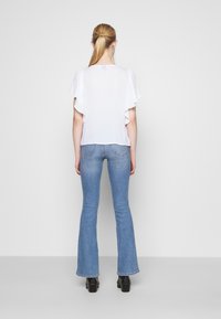 Top bianco a maniche corte con bordi arricciati, abbinato a jeans a campana blu chiaro e stivali neri con tacco, visto da dietro.