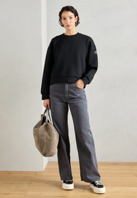 Donna in piedi all'interno che indossa una felpa nera e pantaloni larghi grigi, con una borsa a tracolla beige, con una mano in tasca.