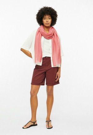 Femme debout de face portant une chemise blanche, un foulard rose, un short bordeaux et des sandales noires sur fond blanc uni.