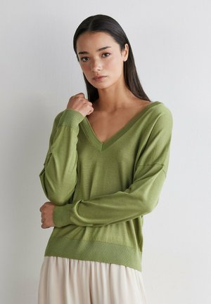 Frau mit langem dunklem Haar, die einen hellgrünen V-Ausschnitt-Pullover und einen cremefarbenen Rock trägt, posiert mit einer Hand nahe am Hals vor einer schlichten Wand.