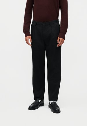 Mężczyzna ubrany w czarne dopasowane spodnie, czarne mokasyny z białymi skarpetkami i ciemnoczerwony, teksturowany sweter z długim rękawem, stojący.