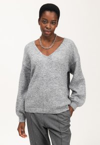Donna che indossa un ampio maglione grigio a scollatura a V, una collana a catena e pantaloni grigi a righe sottili con pois bianchi, in posa con una mano in tasca.